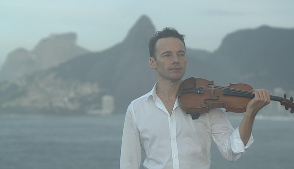 Geiger Linus Roth mit Violine in der Hand und mit weißem Hemd am Strand mit bergen im Hintergrund